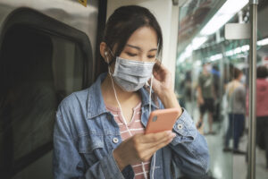 Woman wears a mask, uses a smartphone and earphones, and maintains social distancing when commuting on the subway or train.