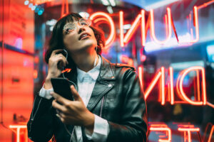 Woman wearing a leather jacket and glasses listens to music against a neon background.