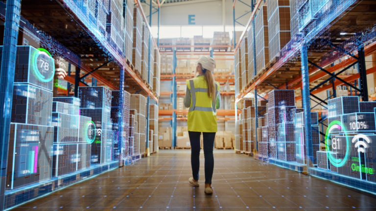 Young woman in high visibility vest and hard hat walks through a warehouse of boxes.