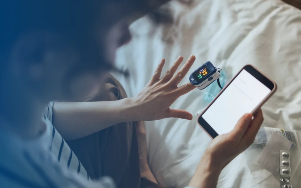 A person looking at their hand with a pulse oximeter attached to one finger and a smartphone.