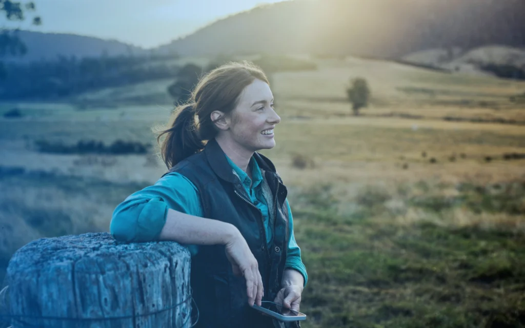 Smiling young woman with a smartphone in her hand leans on a wooden fencepost and looks out at a rural setting.