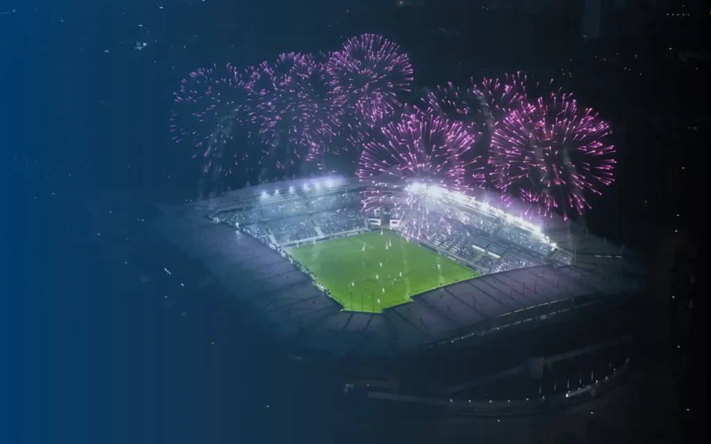 Illuminated sports stadium at night with fireworks above.