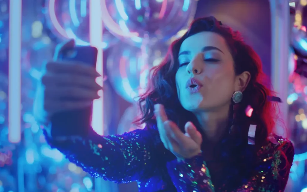 Young woman dressed in sequins blows kisses to a smartphone.