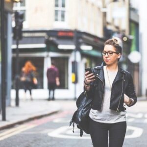 Young woman in leather jacket and glasses looks at mobile phone while walking down the street.