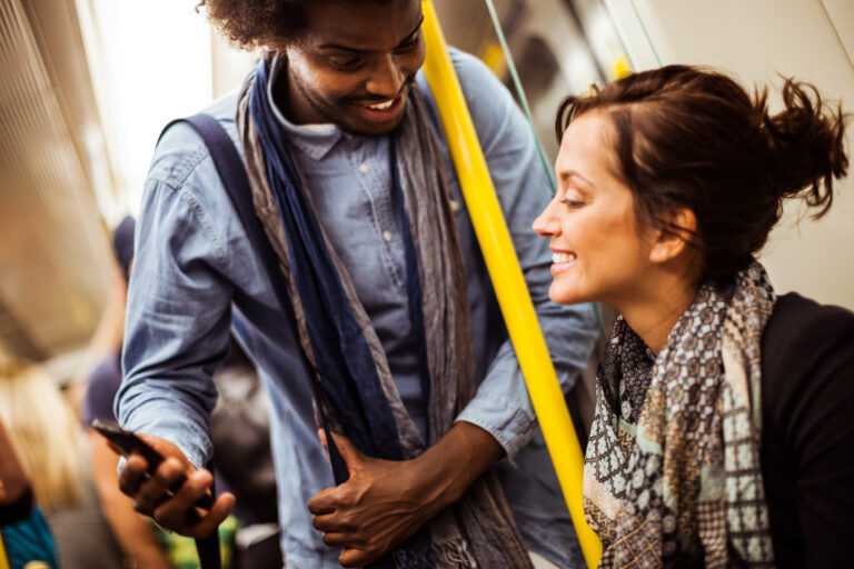 On a train a standing young man show's seated young woman his phone screen.