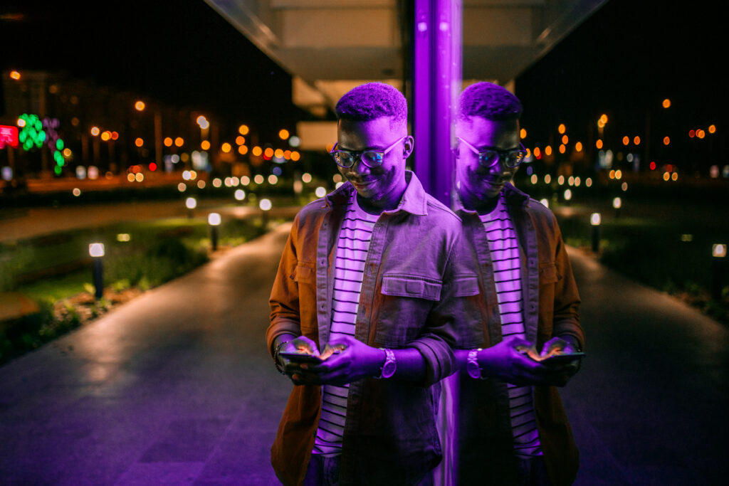 Young man using phone, typing text message in a front of neon blue light office window in rainy night.